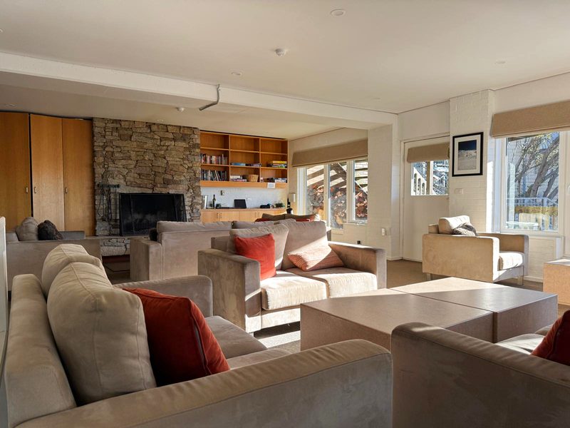 The mid-level lounge: deep sofas and armchairs drawn up to a stone open fire, shelves of books and board games alongside, and windows looking out to the stacked woodpile.